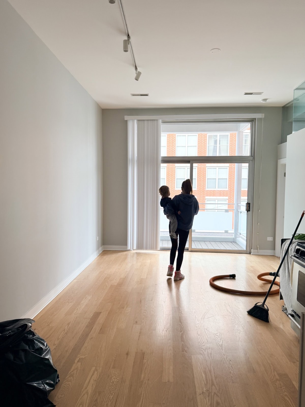 A person holding a child stands in an empty apartment, starting holiday season planning amid cleaning supplies on the floor.