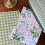 Perfect for shopping organization: green gingham paper, floral folder, and a zipper pouch sit on a desk with a brass lamp.