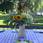 A white pitcher with fresh flowers sits on a blue gingham tablecloth, showcasing the latest in sunny garden dining style.