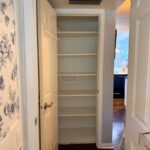 Open closet with five empty white shelves, latest floral wallpaper, and kitchen visible in the background.
