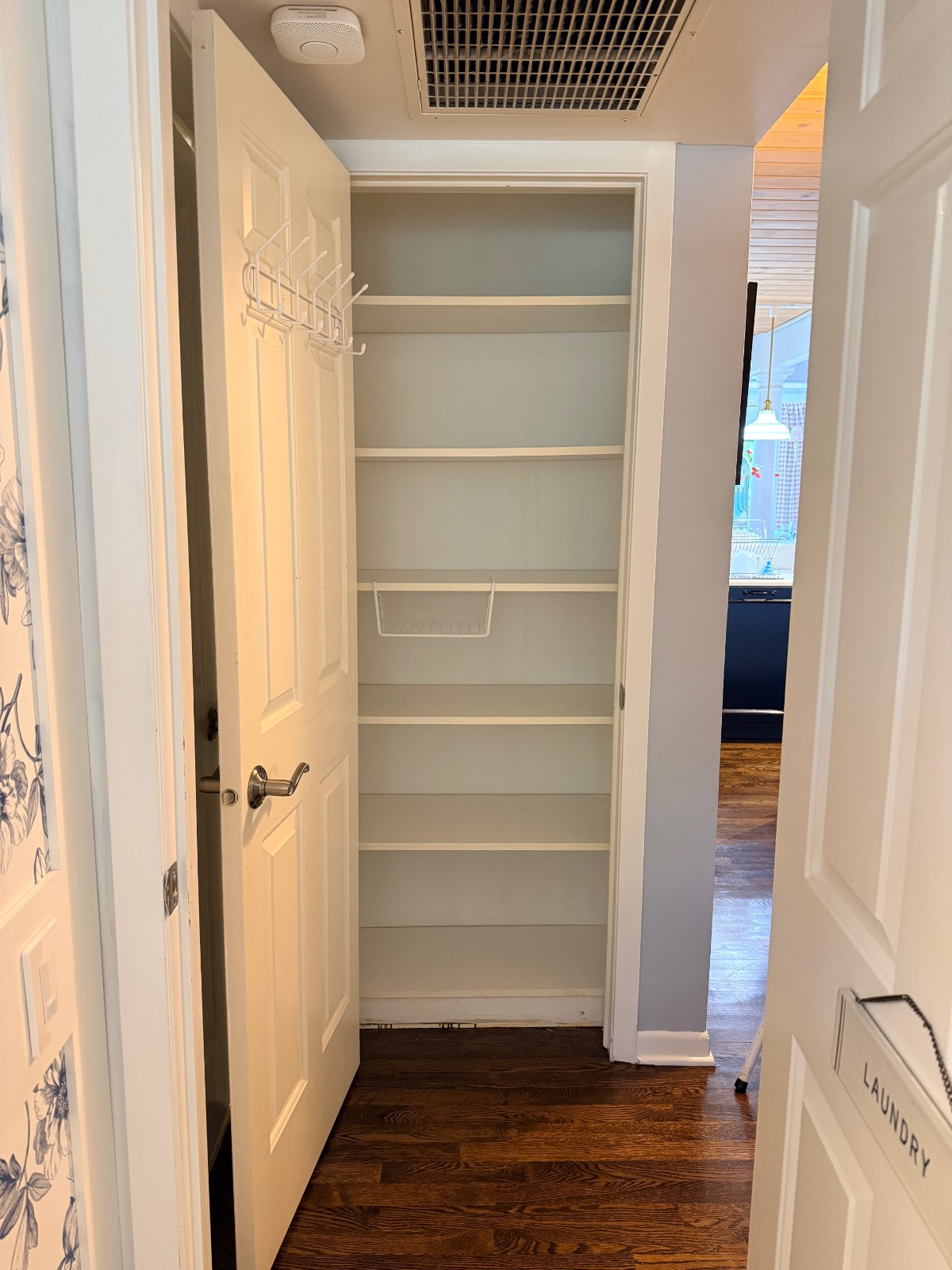 Open pantry with white shelves, door rack, and wire basket adds stylish home organization to dark wood floors.