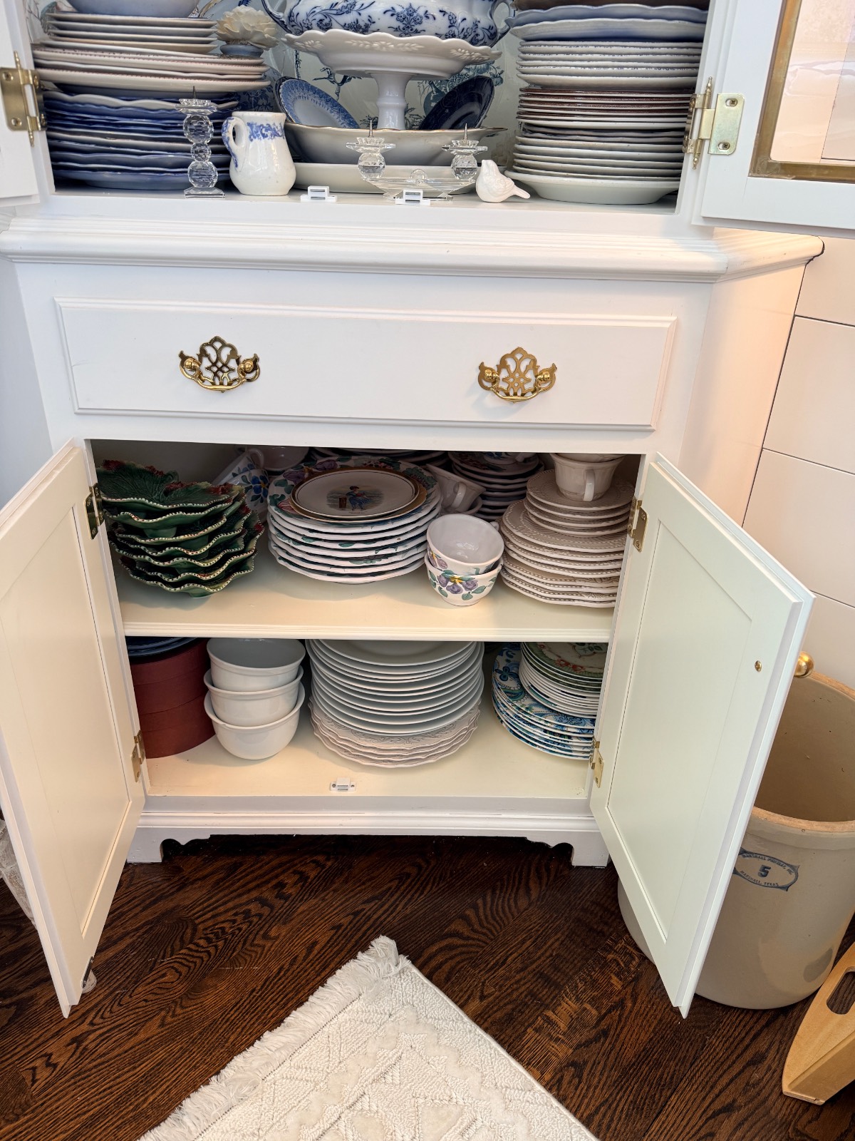 A white hosting closet with stacked plates, bowls, and platters sits on a dark wood floor.