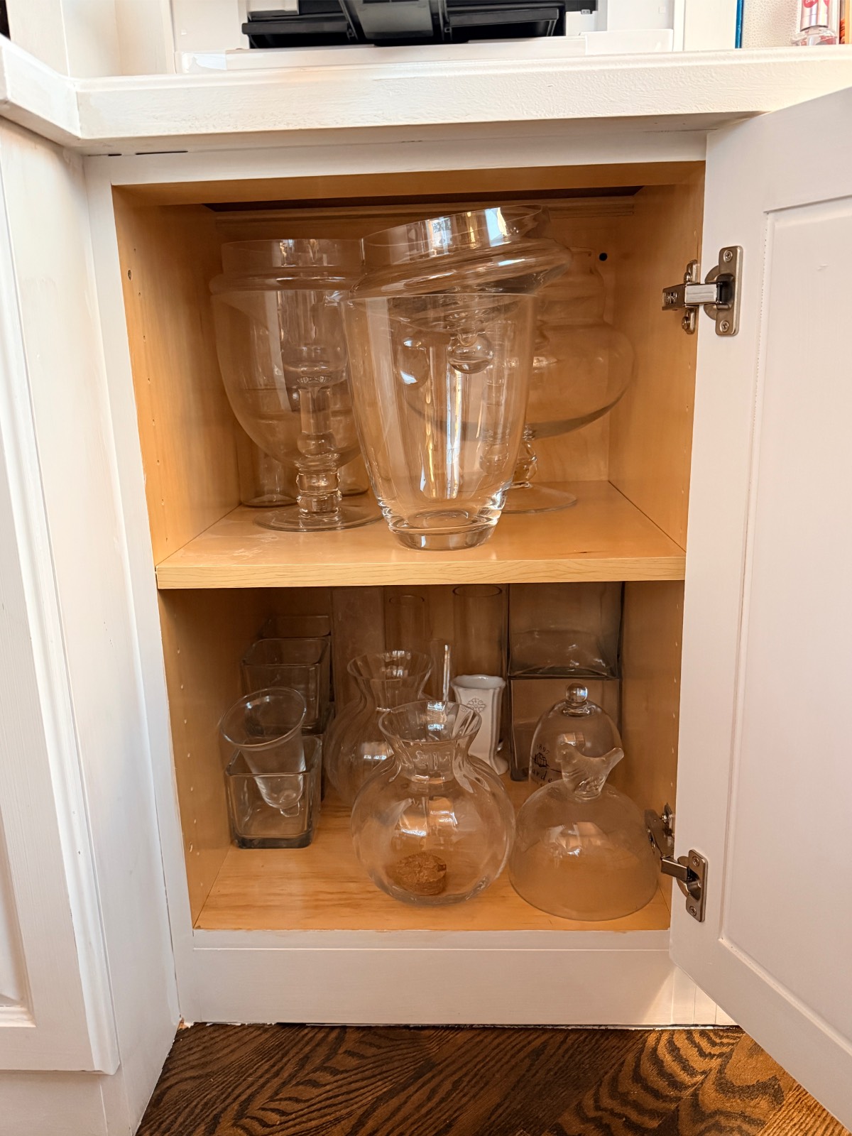 Clear glass vases and pitchers sit on two wooden shelves, creating a stylish hosting closet in an open white cabinet.