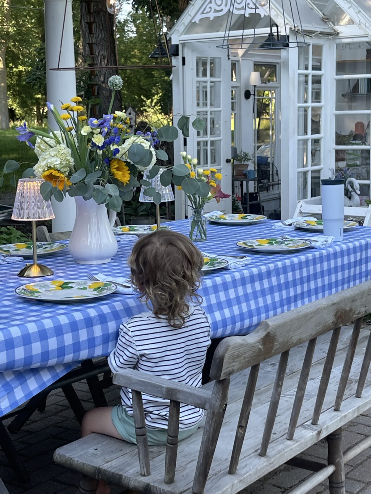 A child sits on a wooden bench at a blue checkered table set with the latest plates, flowers, and lamps.