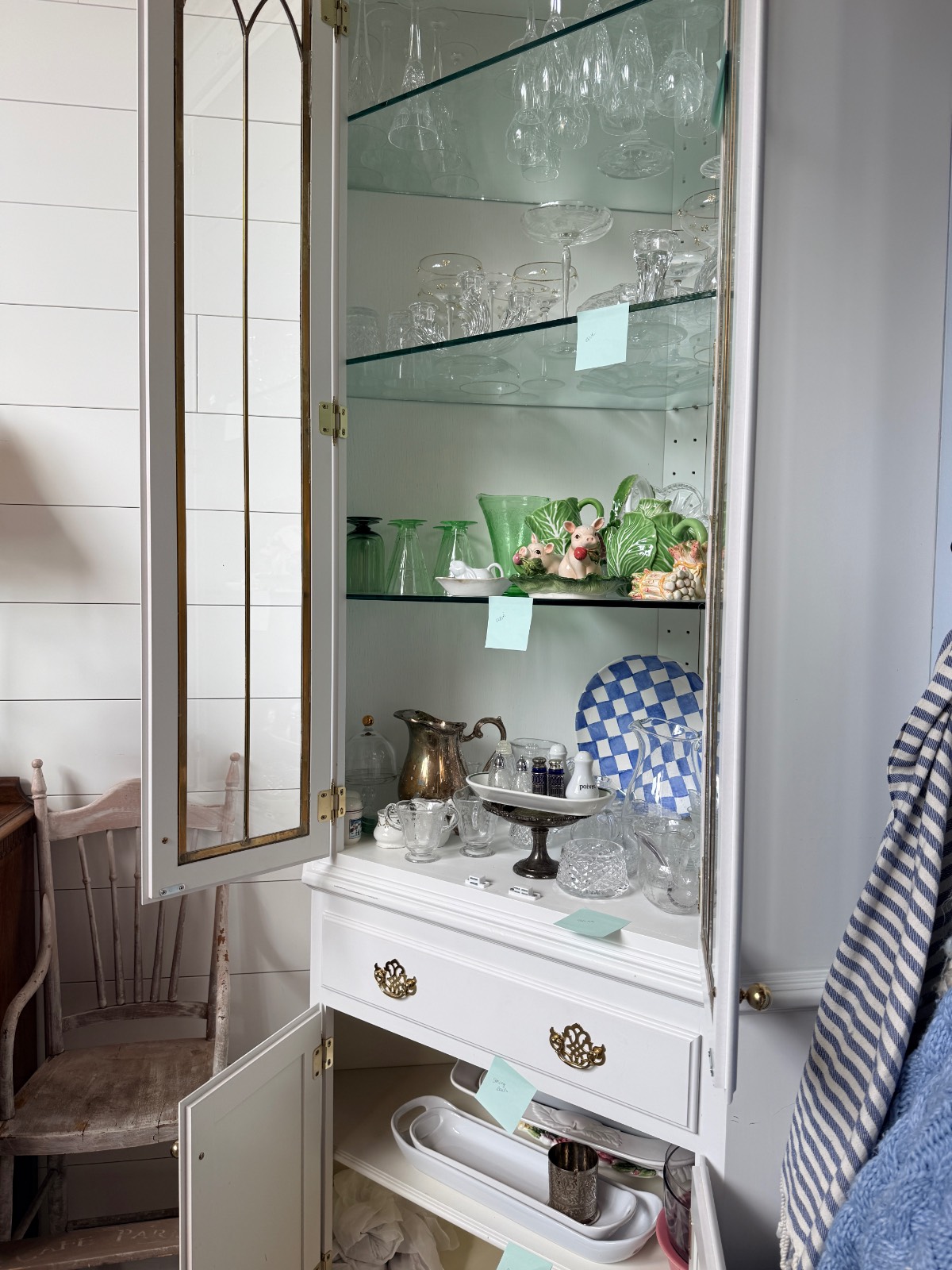 A white cabinet serves as a stylish hosting closet, displaying glassware, a teapot, cake stand, and animal figurines.