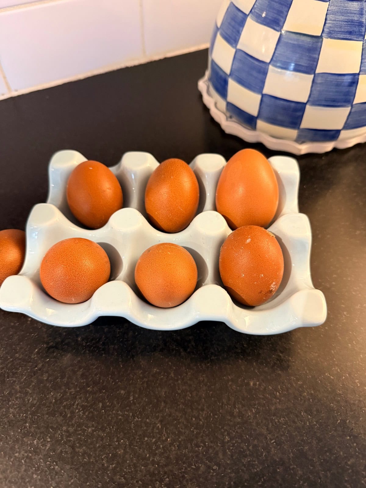 Six brown eggs in a white ceramic egg holder, next to the latest blue checkered container on a dark counter.