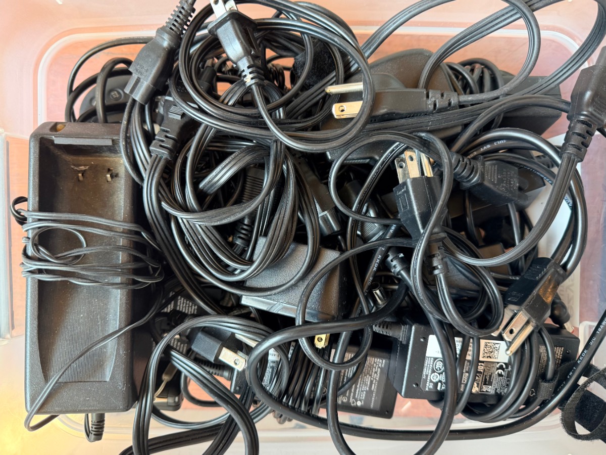 A plastic bin filled with tangled black power cords, chargers, and plugs—things to throw away right now.