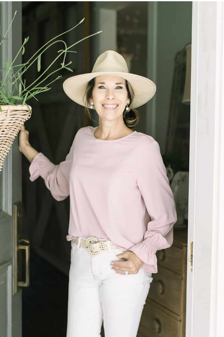 Woman in a straw hat and pink blouse stands smiling in a doorway, showing off her latest casual look.