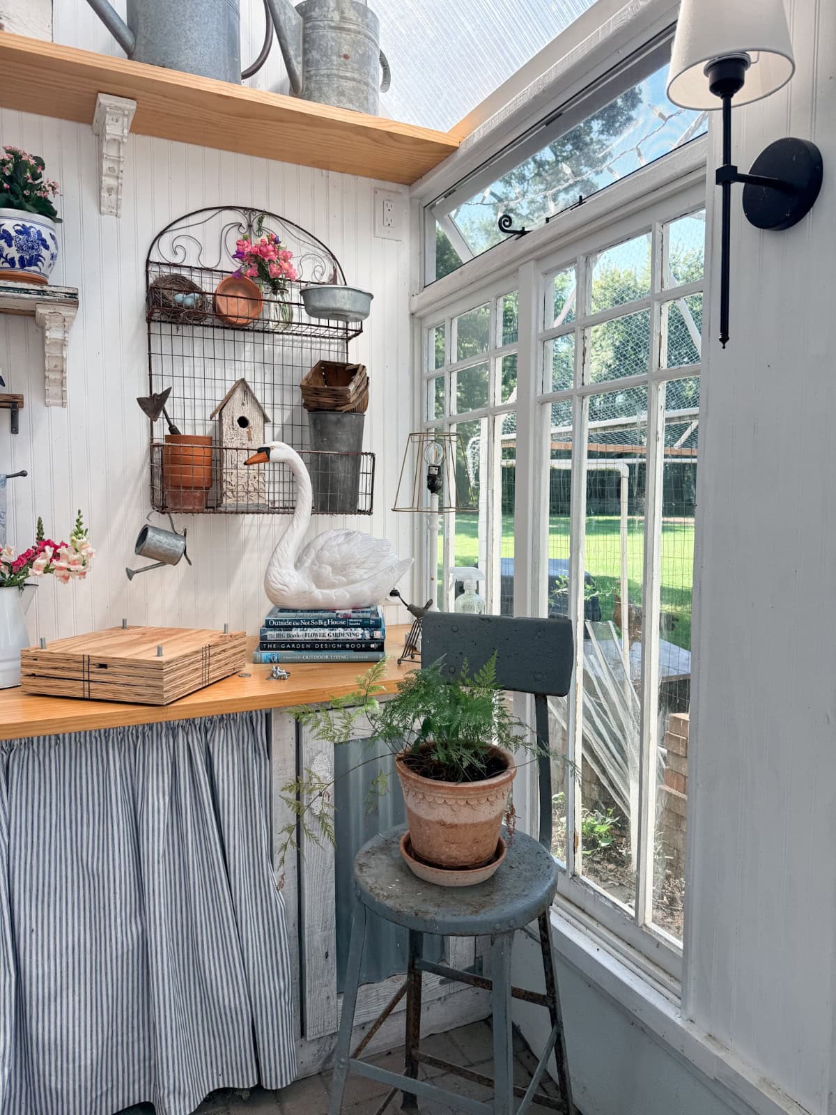 Sunlit garden room with summer styling, potted fern on stool, swan decor, vintage finds, and tools near large window.