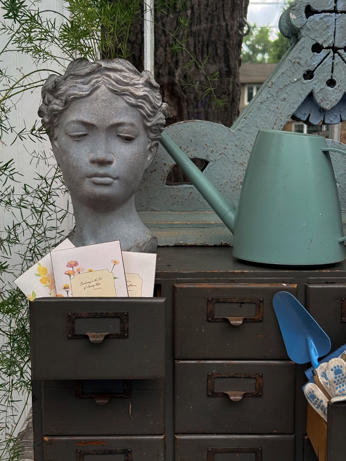 Bust planter, green watering can, and vintage finds styled on drawers with a greenhouse garden backdrop.