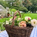 A woven basket of flowers, styled with the latest trends, sits on a blue checked tablecloth in a lush garden setting.