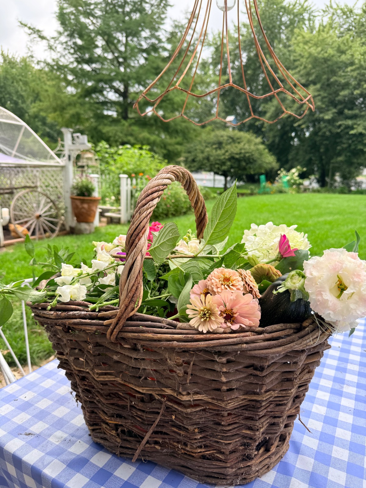 A woven basket of flowers, styled with the latest trends, sits on a blue checked tablecloth in a lush garden setting.