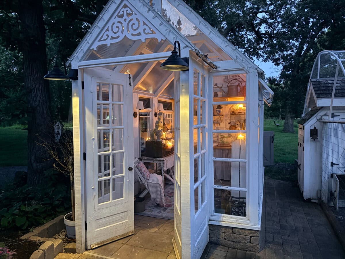 A small, white greenhouse with open doors glows warmly, showcasing vintage finds in the early evening.