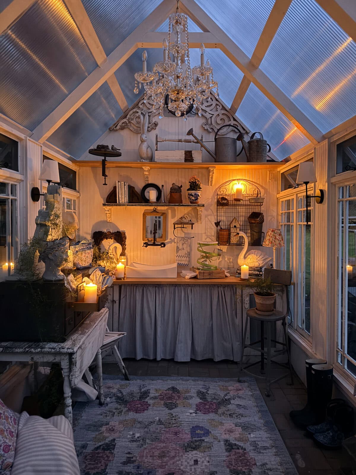 A cozy, candlelit greenhouse with vintage finds, plants, a rug, shelves, and a chandelier hanging from the ceiling.