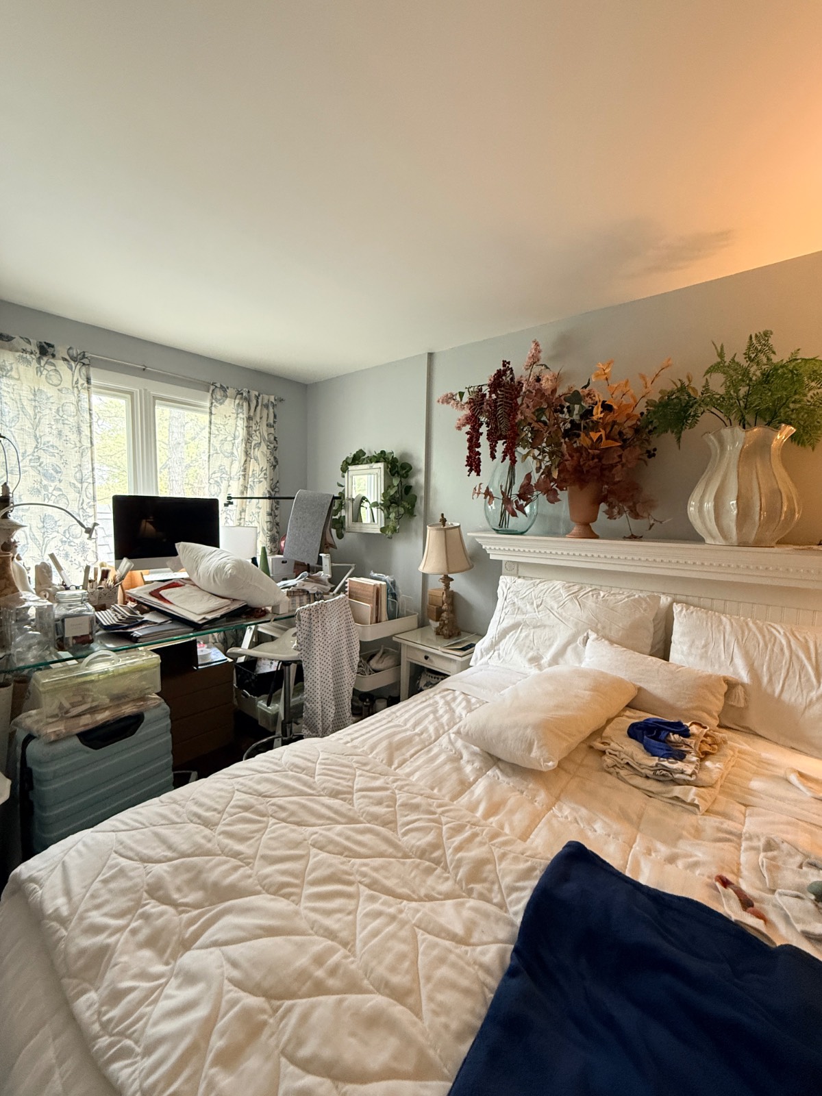 A cozy, cluttered third bedroom with a messy bed, computer desk, plants, and sunlight streaming through the window.