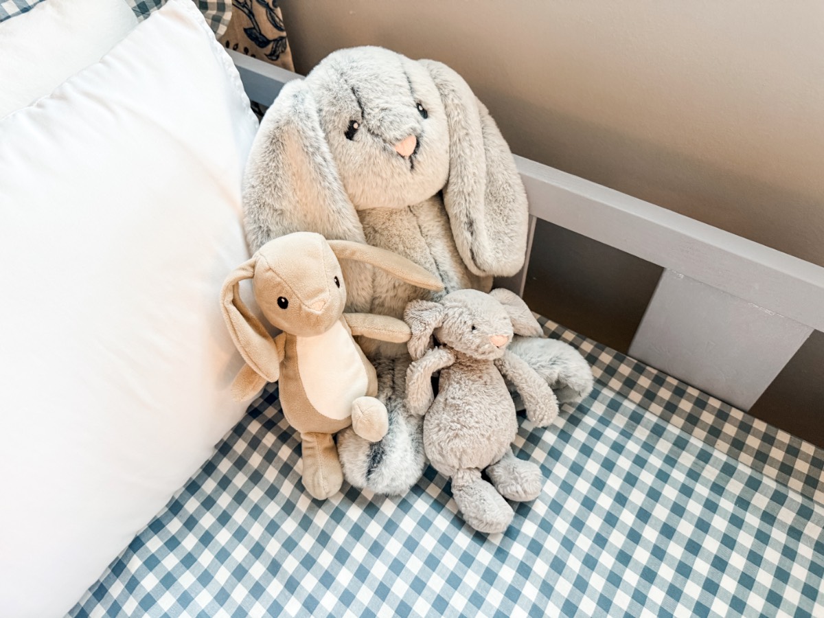 Three plush bunny toys sit together on a cozy, blue and white checkered bed beside a white pillow.