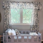 Cozy toddler bed with pillows and stuffed animals sits under a window with blue floral curtains on a sunny day.