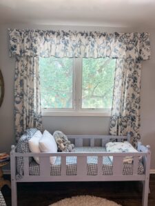Cozy toddler bed with pillows and stuffed animals sits under a window with blue floral curtains on a sunny day.