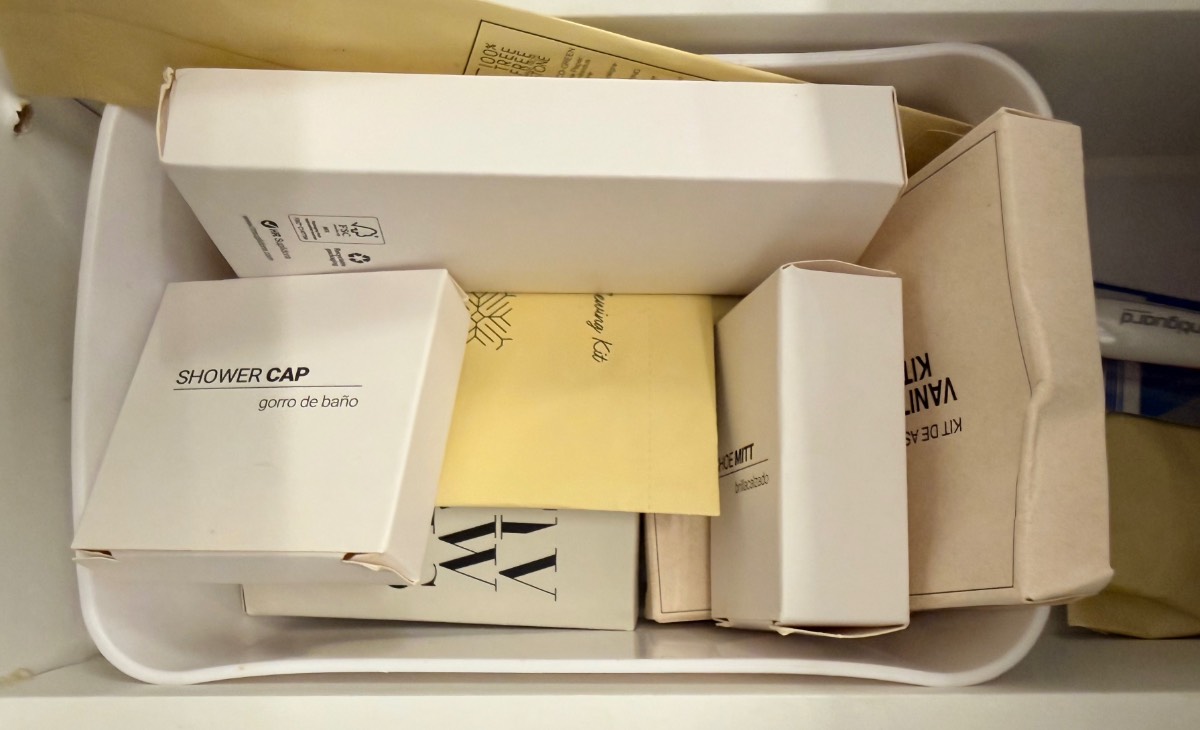 Assorted hotel toiletries—things to throw away right now—like a shower cap and vanity kit, in a white plastic tray.