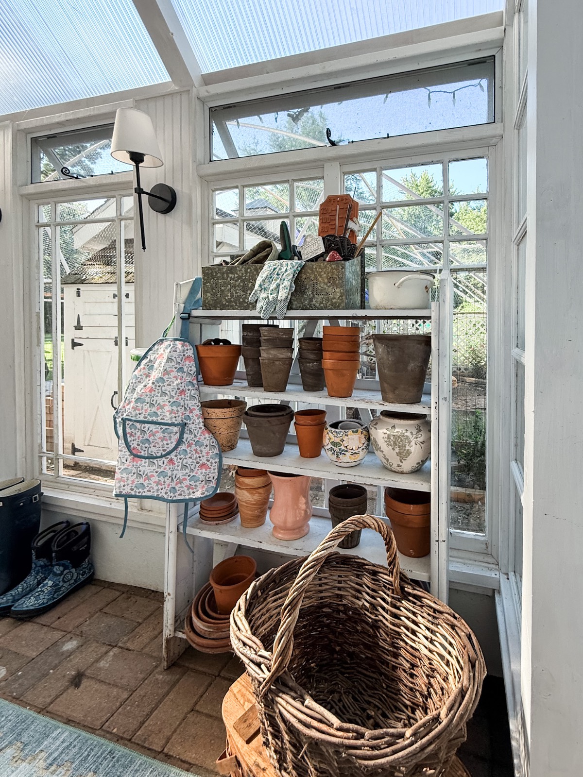 A shelf with empty flower pots, vintage finds, and a floral apron adds charm to this sunlit greenhouse.