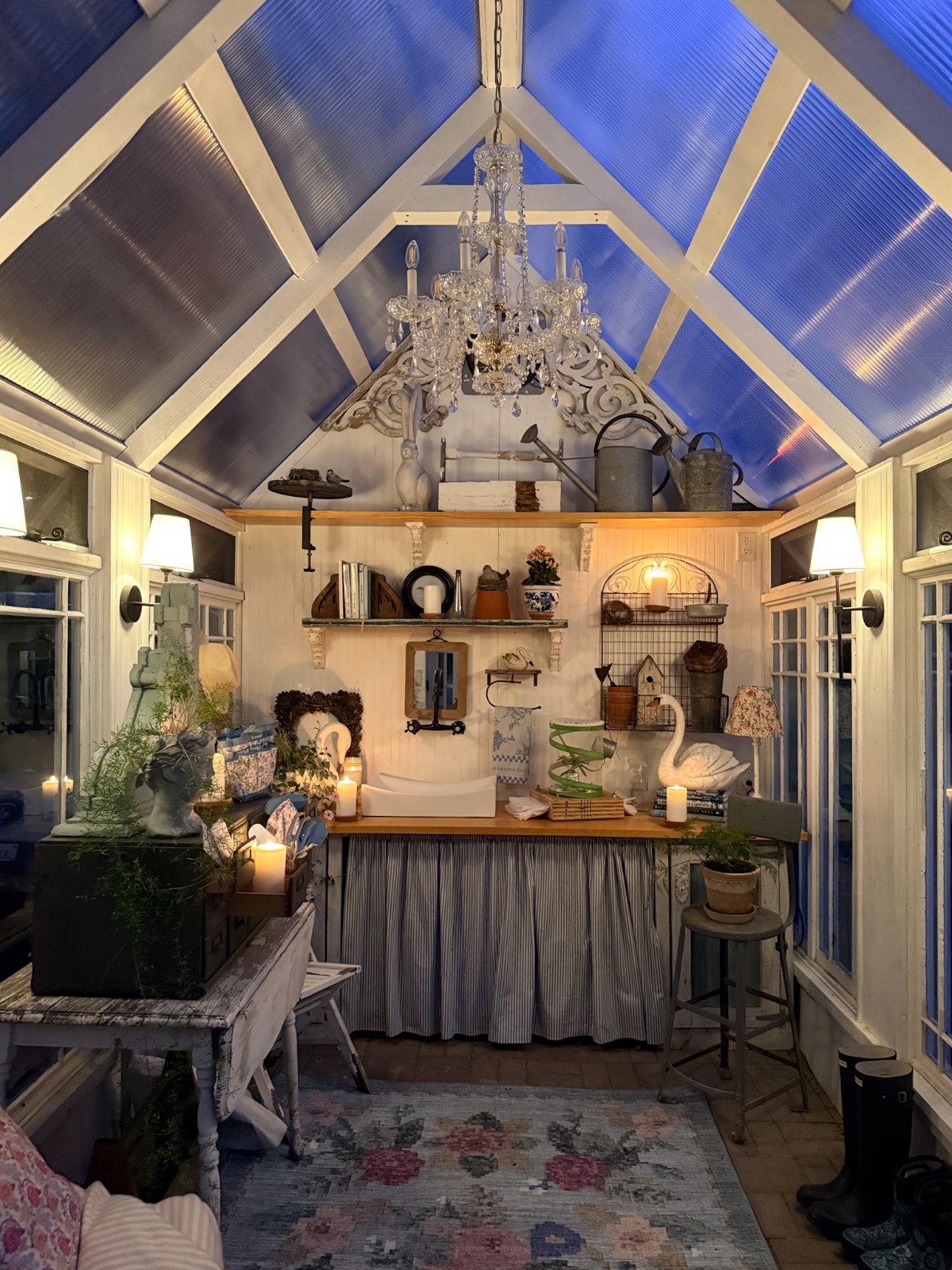A cozy, well-lit greenhouse styled for summer with vintage finds, shelves of plants, a chandelier, and eclectic decor.