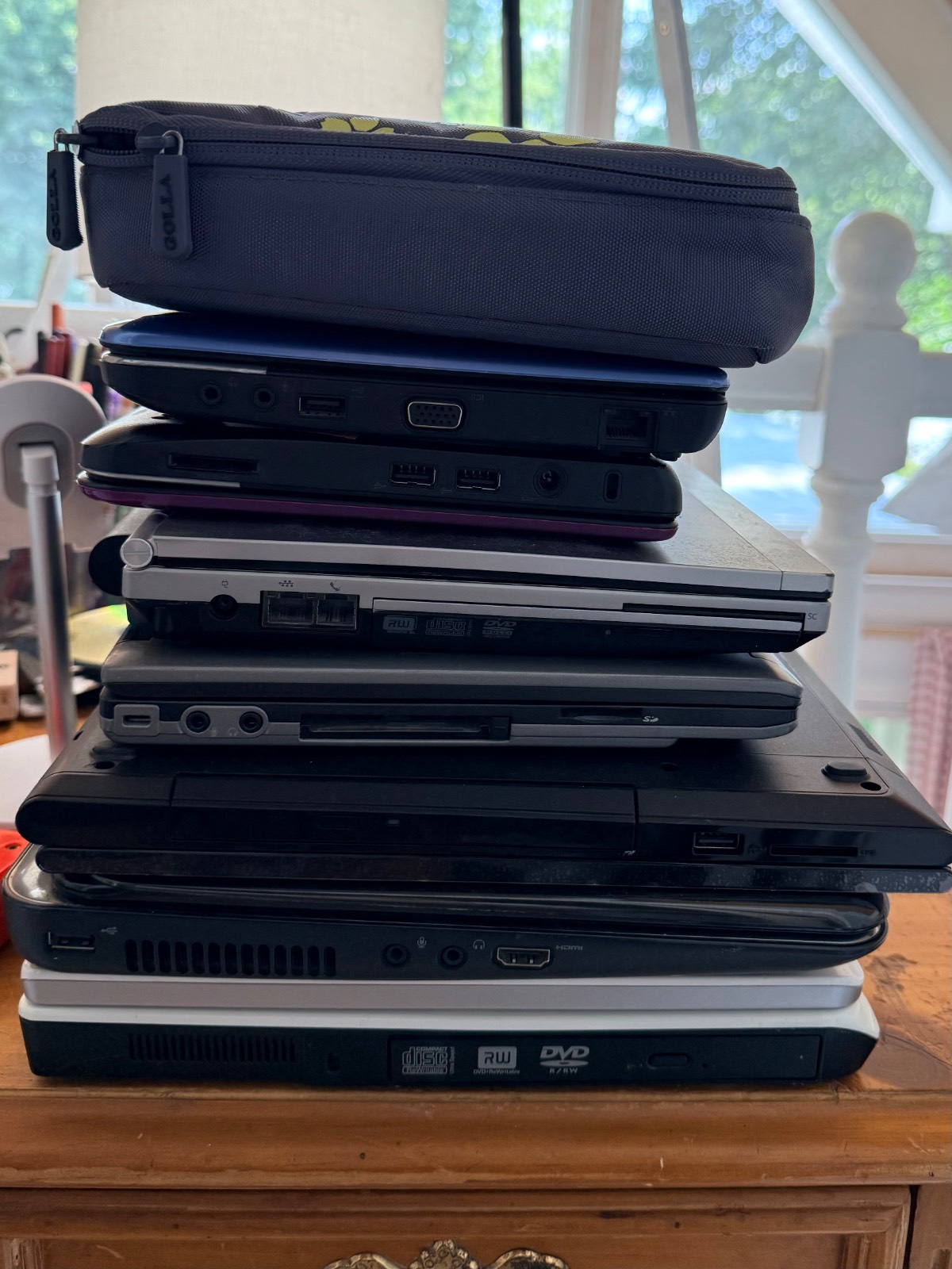 A stack of old laptops—things to throw away right now—and a pencil case sit on a wooden desk by a window with greenery.