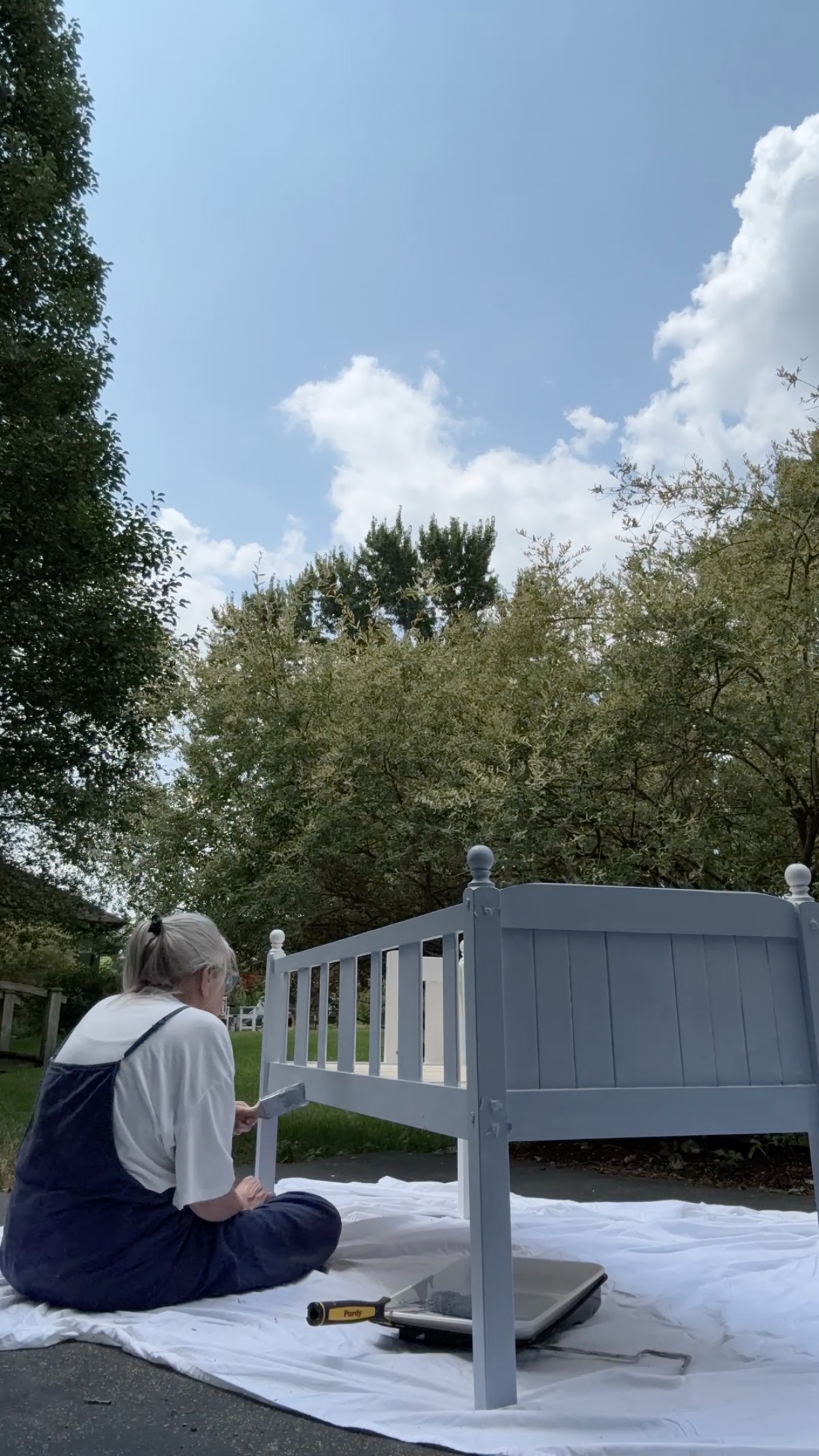 A person paints a blue wooden bed frame for a cozy family suite outdoors on a white tarp under a partly cloudy sky.
