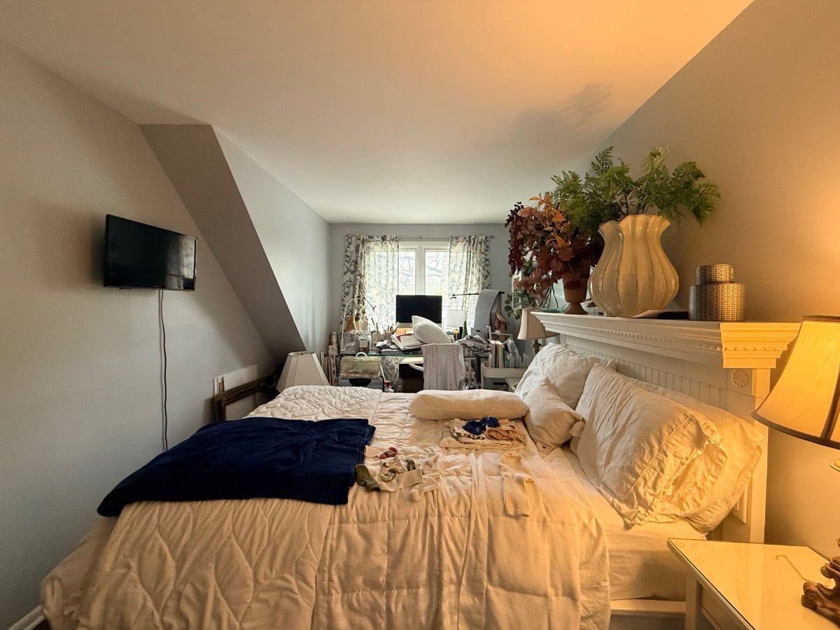A messy room with a bed, wall-mounted TV, desk by the window, and plants on a white mantel above the bed.