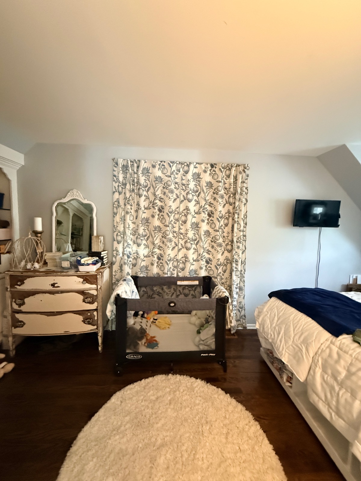 Cozy third bedroom with crib, dresser, mirror, wall TV, patterned curtains, and a bed with blue blanket.