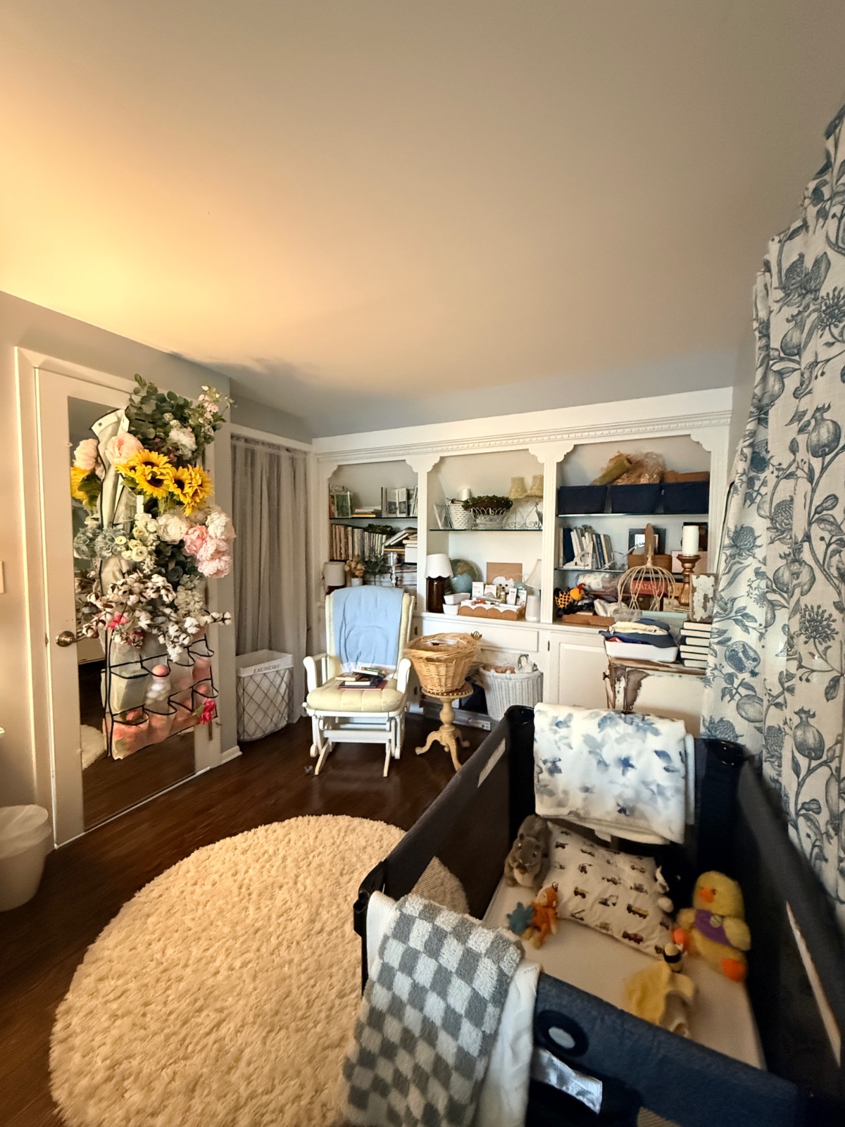 Cozy nursery in the third bedroom with crib, armchair, bookshelf, floral decor, toys, and soft lighting.