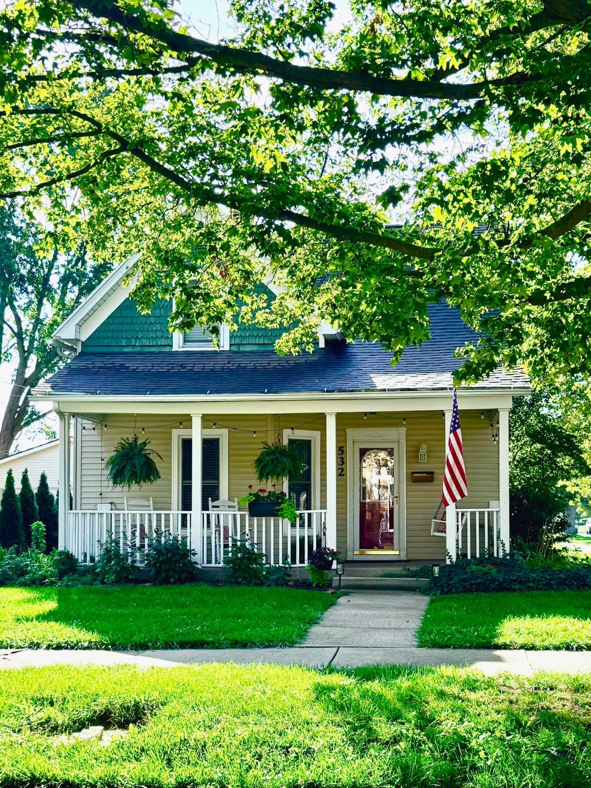 A tan cottage-style house with a porch, hanging plants, and flag—found near the local mercantile market, tree-shaded.