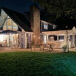A cozy house with a lit patio, outdoor tables, and latest stone chimney at night, surrounded by grass and trees.