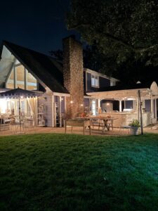 A cozy house with a lit patio, outdoor tables, and latest stone chimney at night, surrounded by grass and trees.