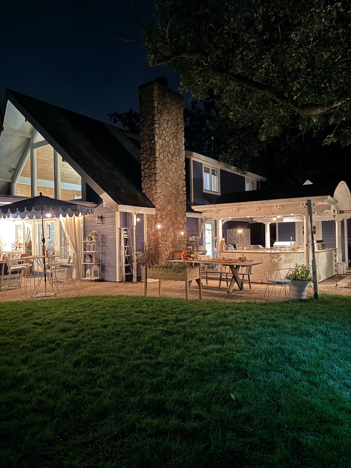 A cozy house with a lit patio, outdoor tables, and latest stone chimney at night, surrounded by grass and trees.