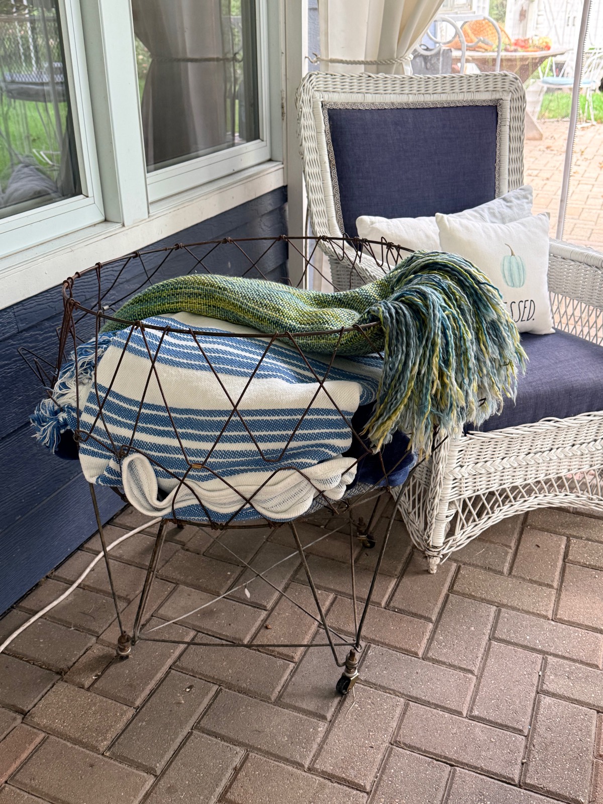 Striped and green blankets in a metal basket add muted fall decor beside a white wicker chair with blue cushions on a patio.