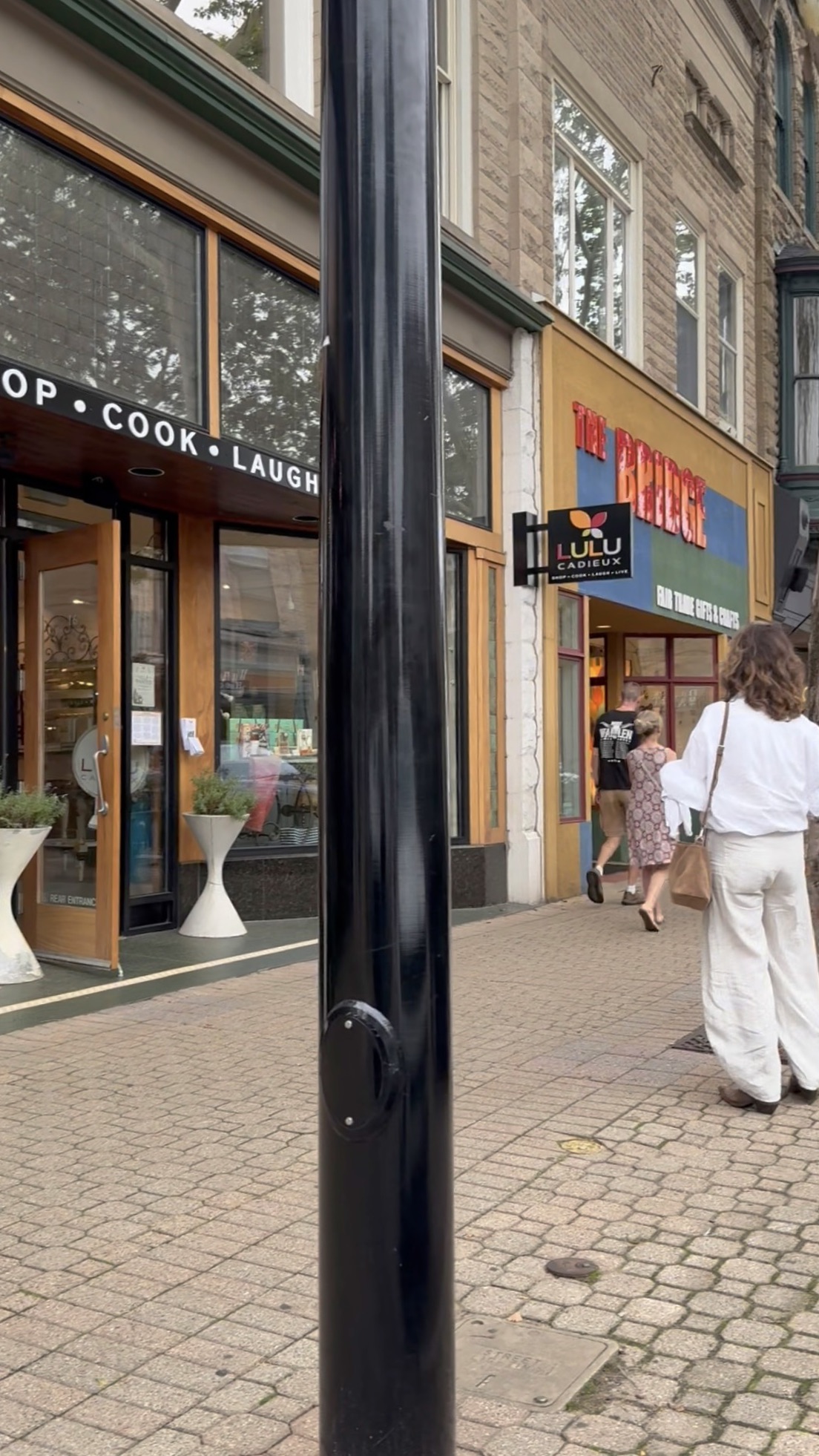 A sidewalk with people walking past shops, including Found Cottage Mercantile market, and a black pole in the center.