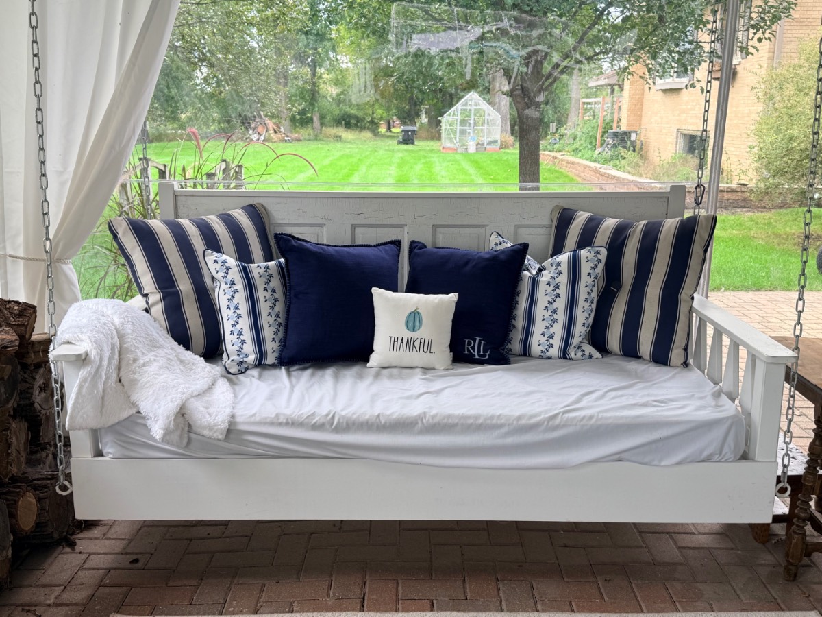 White porch swing with blue and striped pillows, plus a throw, creates cozy muted fall decor amid backyard grass and trees.