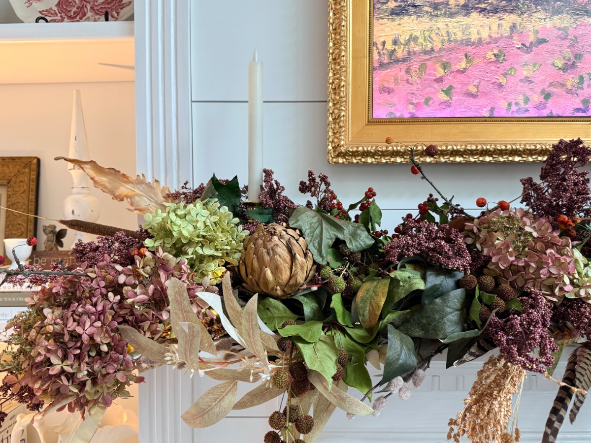 A floral arrangement with hydrangeas, artichoke, berries, and candle adds muted fall decor below a gold-framed painting.