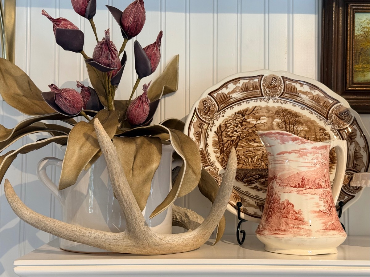 Antler, faux fig arrangement, cranberry platter, and pink pastoral pitcher on a white shelf with paneling.