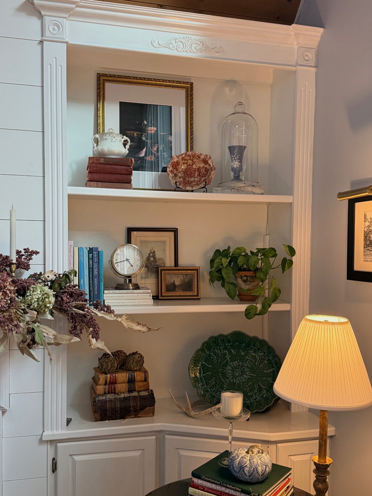 White corner bookshelf with books, muted fall decor, a plant, framed art, and a cranberry-hued lamp softly glowing nearby.