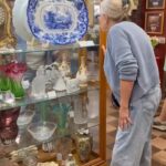 Woman in light blue sweater and jeans browses antiques at a found cottage mercantile market display case.