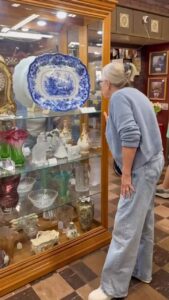 Woman in light blue sweater and jeans browses antiques at a found cottage mercantile market display case.
