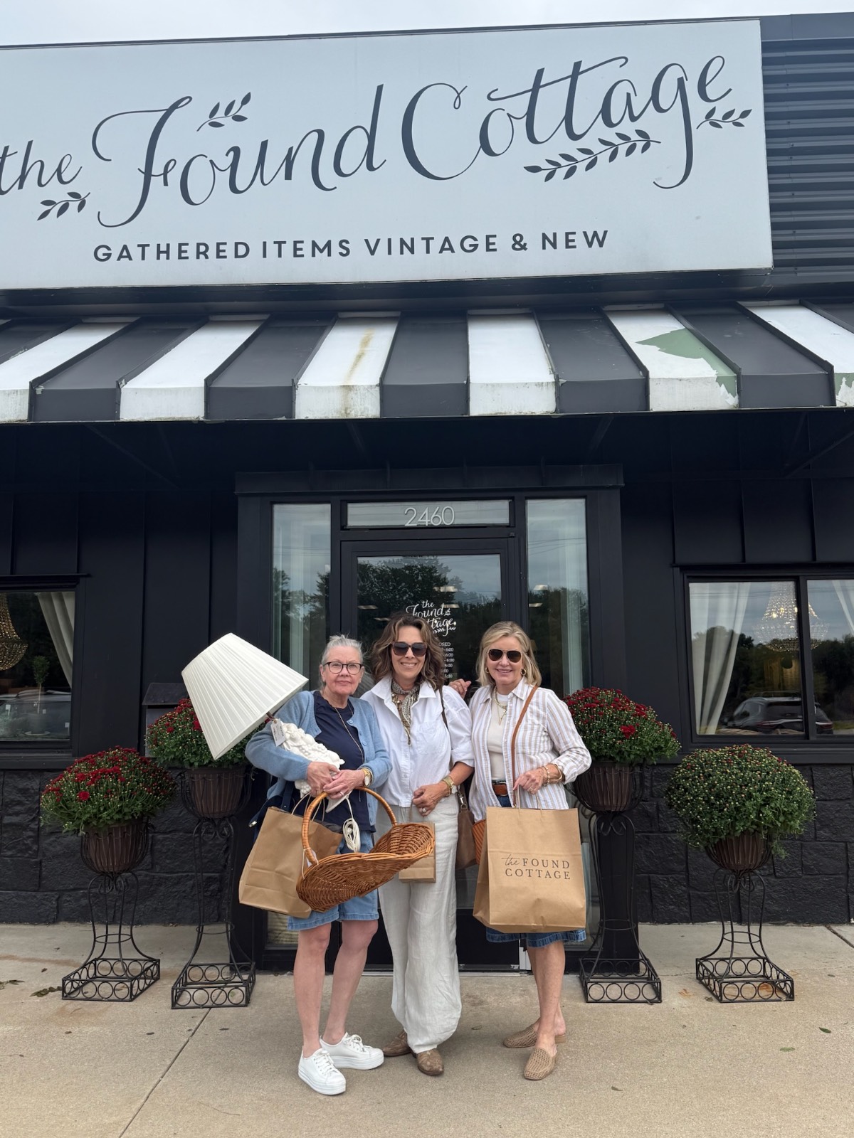 Three women smile outside The Found Cottage Mercantile Market, holding shopping bags and a lamp near potted plants.