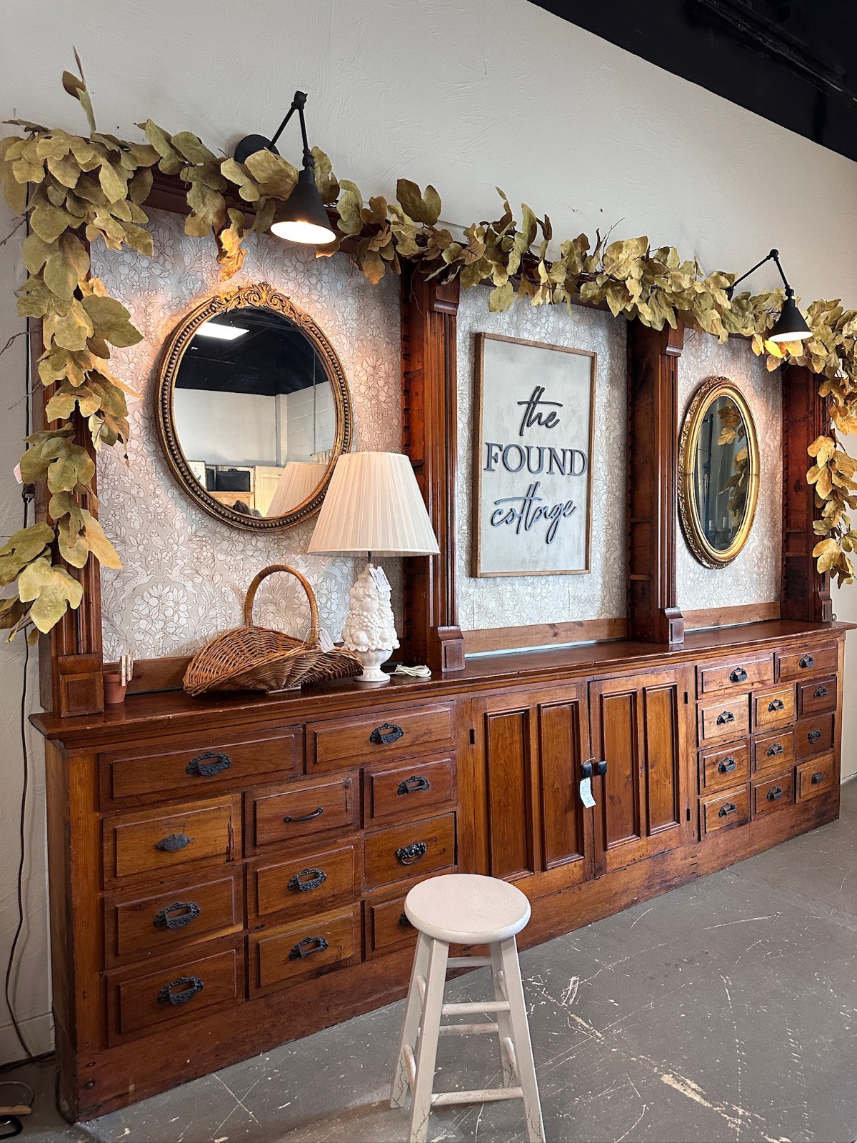 Large wooden cabinet with mirrors, leafy garland, art, lamps, and a stool—found cottage mercantile market charm.