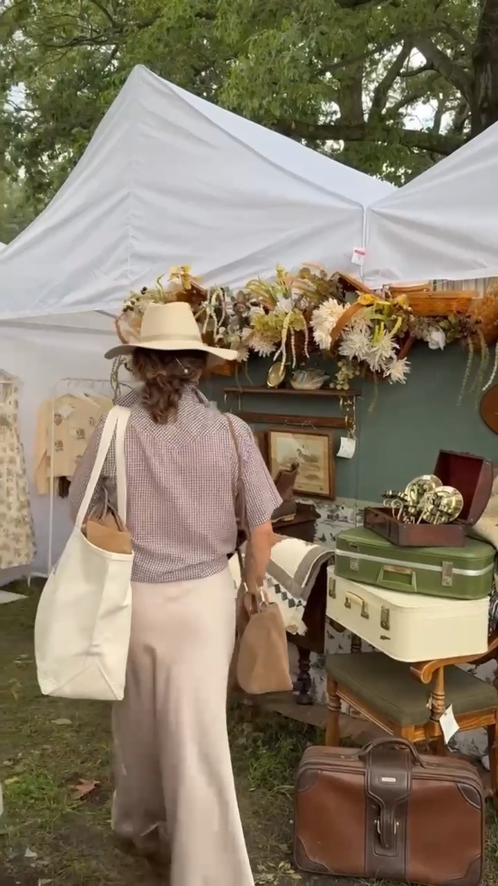 A woman in a hat browses a found cottage mercantile market stall with suitcases, clothes, and floral decorations.