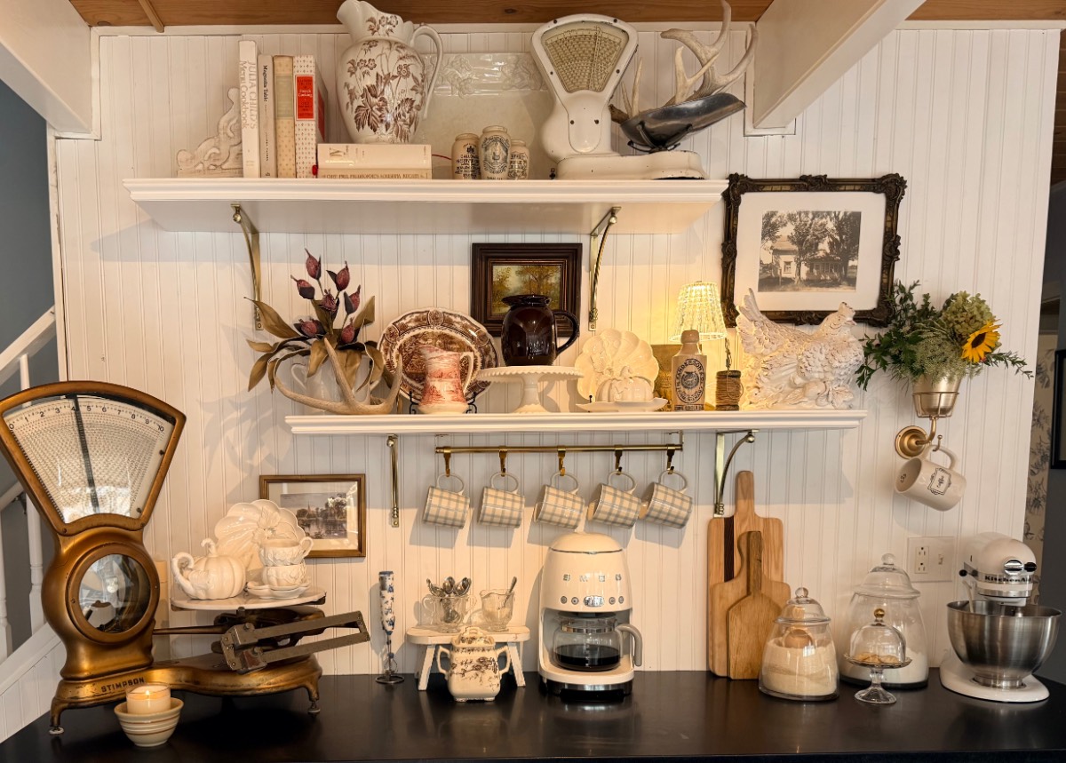 Vintage kitchen shelves with mugs, scales, and jars featuring muted fall decor on a white beadboard wall.