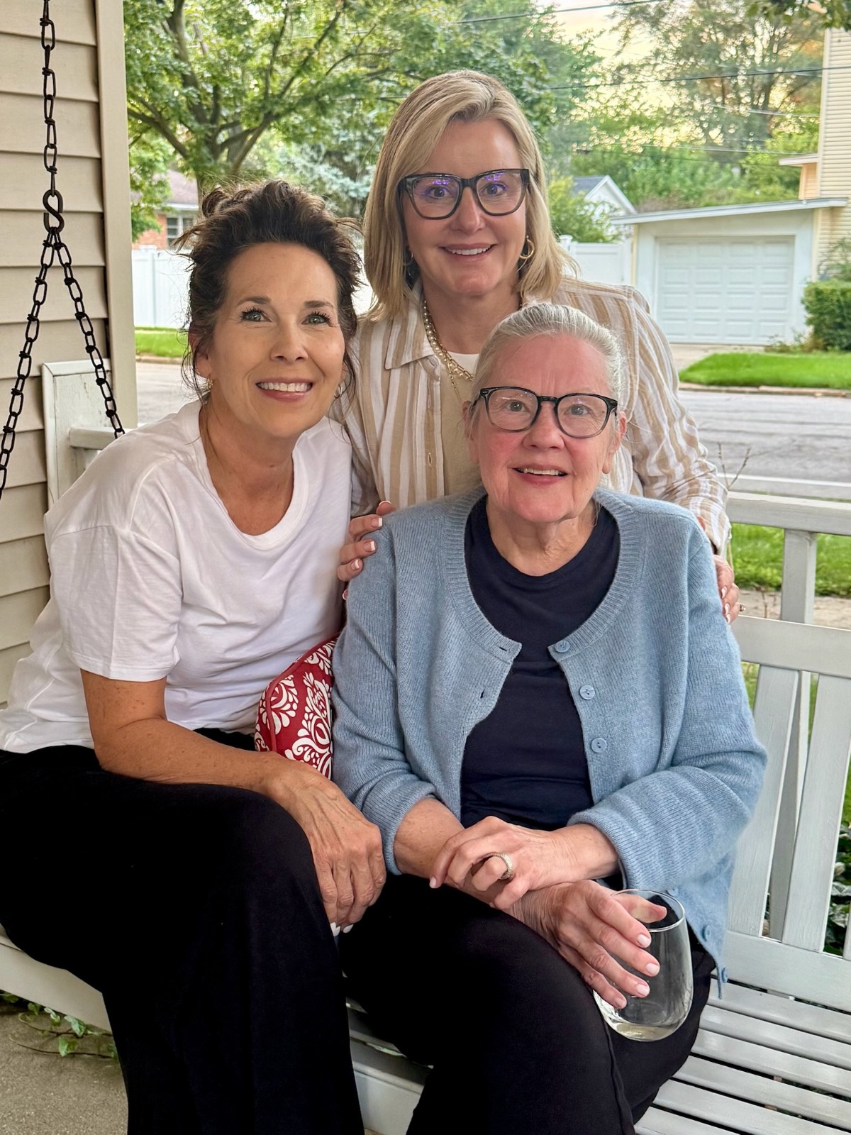 Three smiling women sit together on a porch swing at the found cottage, with lush greenery in the background.