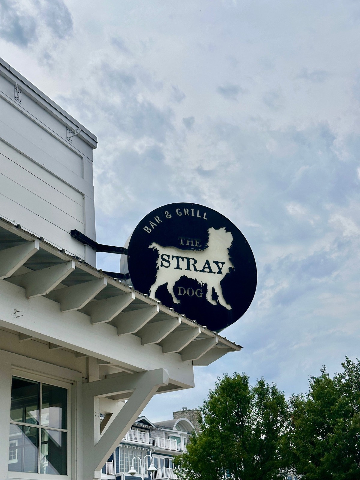 Round sign reads “The Stray Dog Bar & Grill” with a dog silhouette, found on a white building against a cloudy sky.