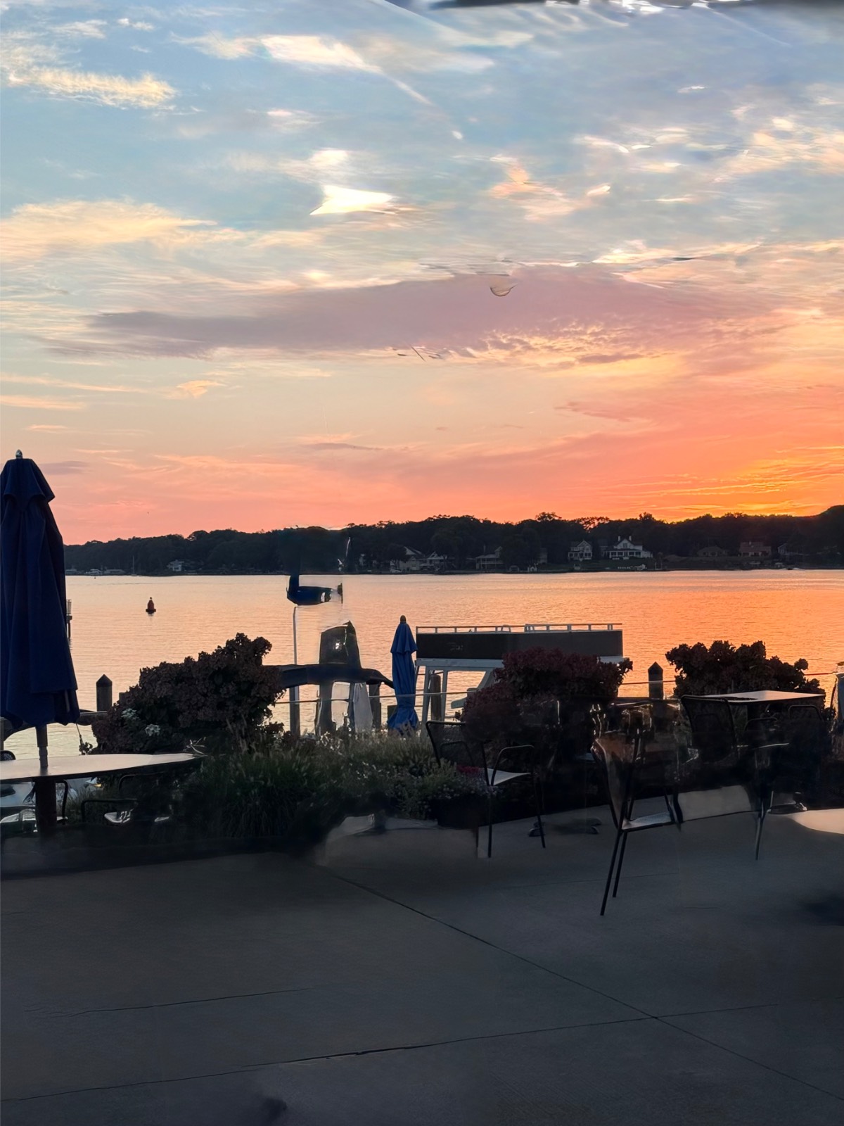 Patio by lake at sunset, with empty tables, chairs, umbrellas, dock, and sky near the Found Cottage Mercantile Market.