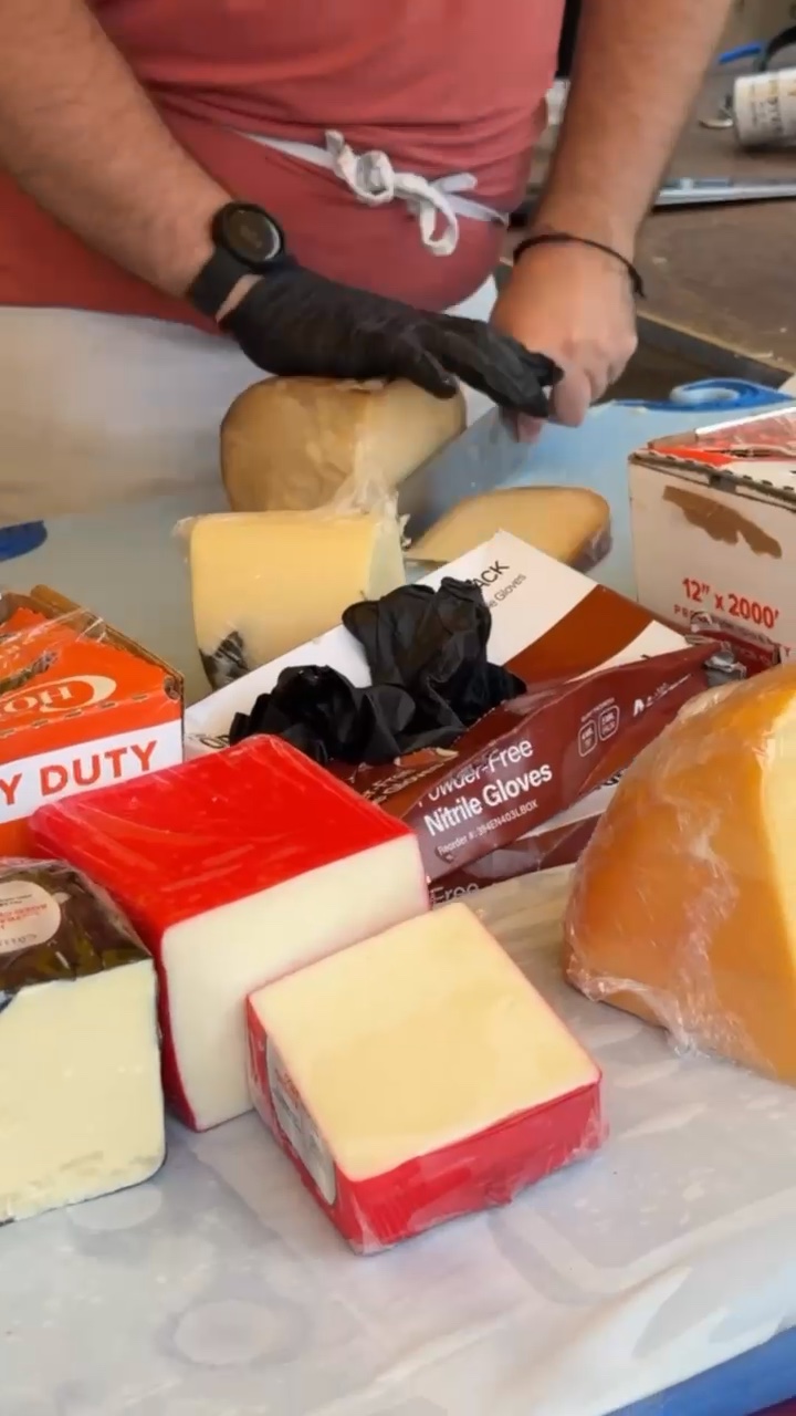 At the Holland Farmers Market, a gloved person slices cheese blocks; assorted cheeses and gloves are arrayed on the table.